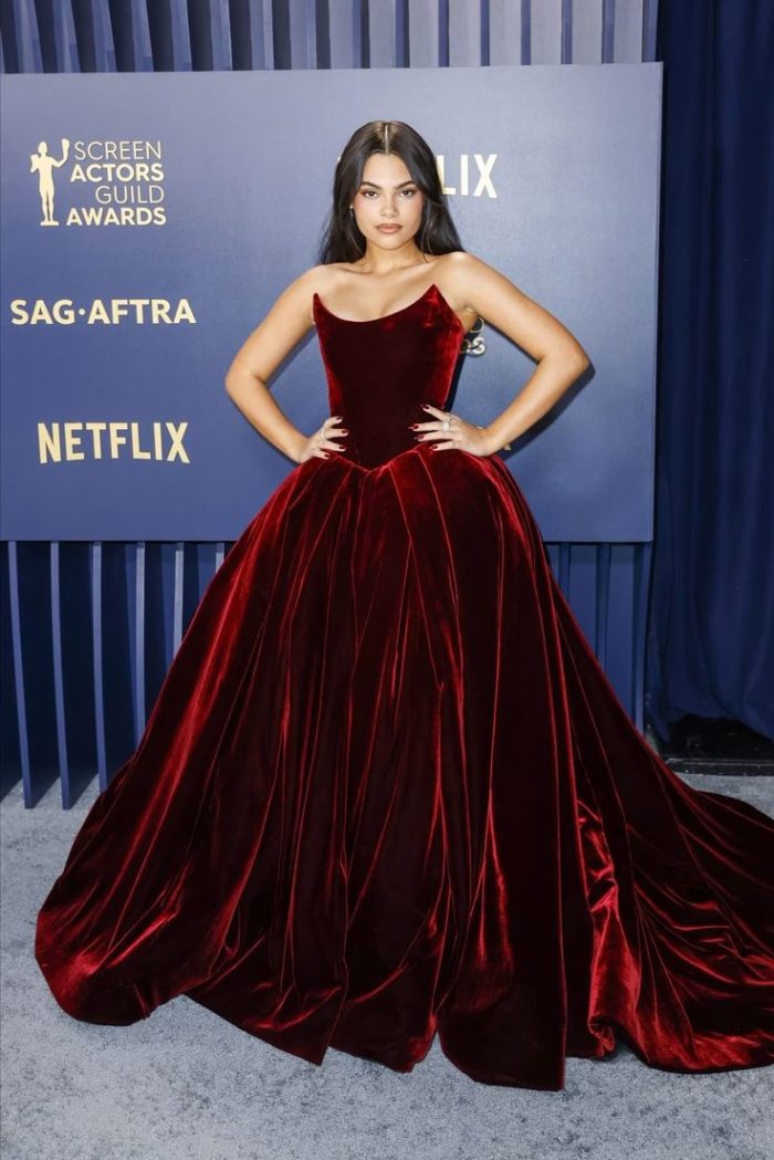 Ariana Greenblatt wearing a Deep red velvet strapless ballgown with a dramatic cat-eye neckline, structured bodice, and full flowing skirt at a formal awards event