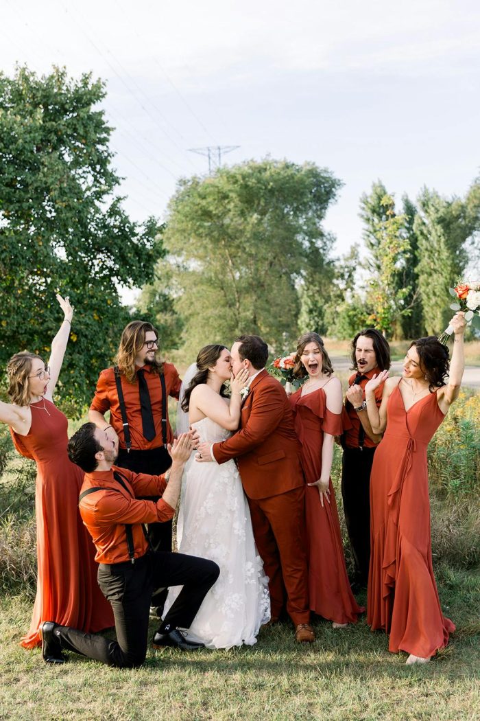Bride wearing the Laila by Maggie Sottero wedding dress celebrates with her wedding party in rust-colored attire during an outdoor wedding