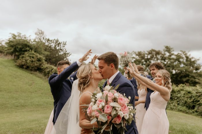Bride wearing Fontaine by Maggie Sottero kisses her husband in front of bridesmaids and groomsmen