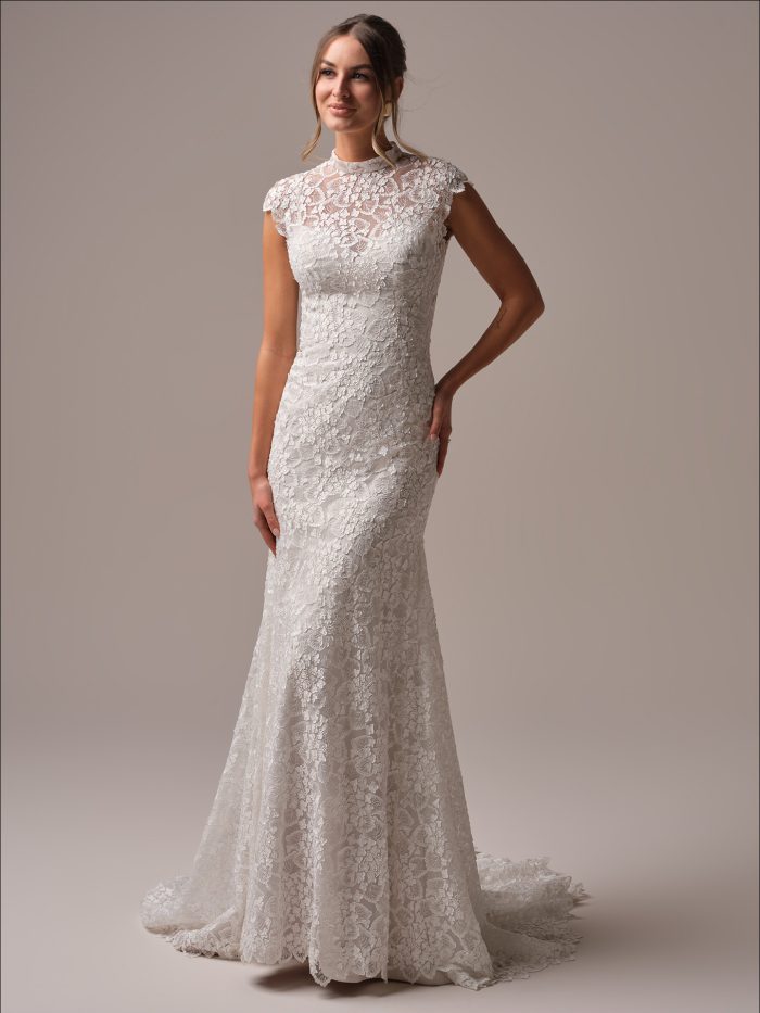Bride wearing a modest sheath wedding dress with allover floral lace, high illusion lace neckline, and cap sleeves, standing in a studio with train displayed.