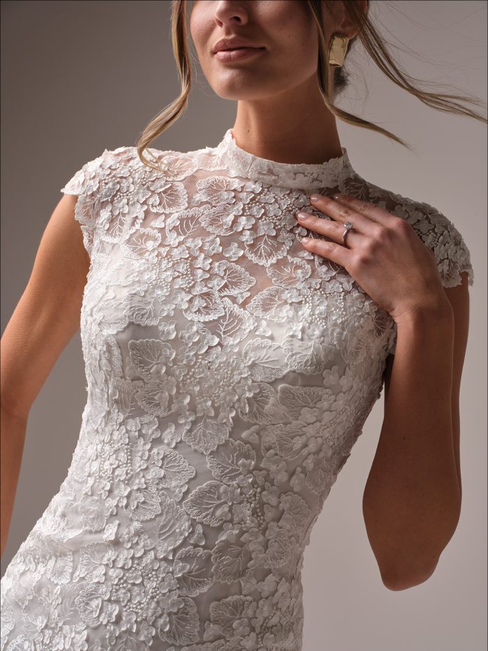 Close-up of a modest sheath wedding dress bodice showing intricate floral lace appliqué, high illusion neckline, and delicate cap sleeve detail.
