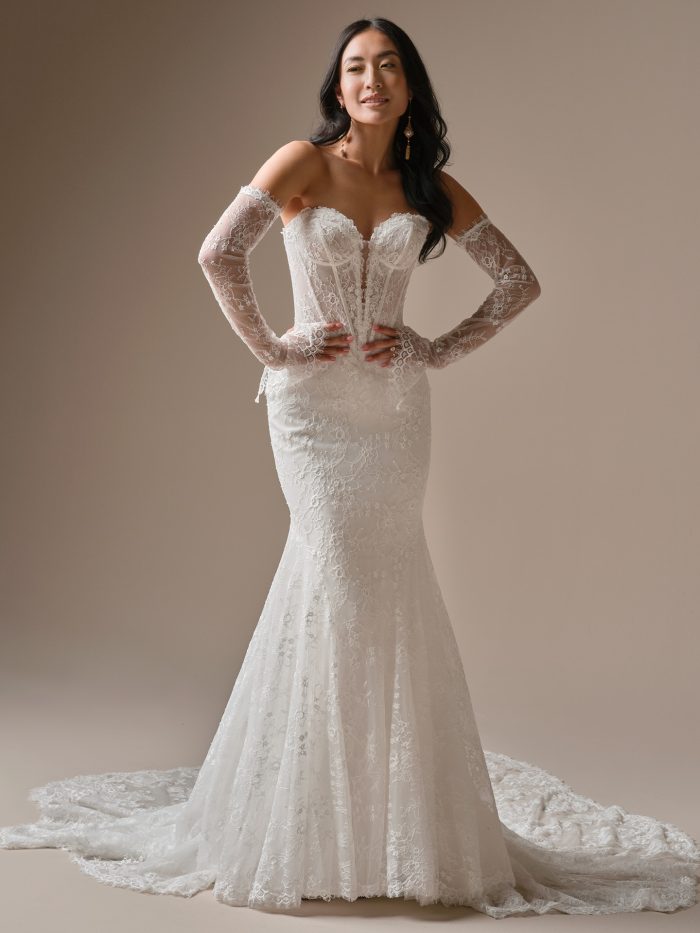 Bellevue wedding dress featuring unembellished Chantilly lace, a strapless sweetheart deep illusion neckline, exposed boning along the illusion bodice, and a fit-and-flare silhouette with an extended scalloped lace train. Model stands with hands on hips wearing matching lace arm sleeves.