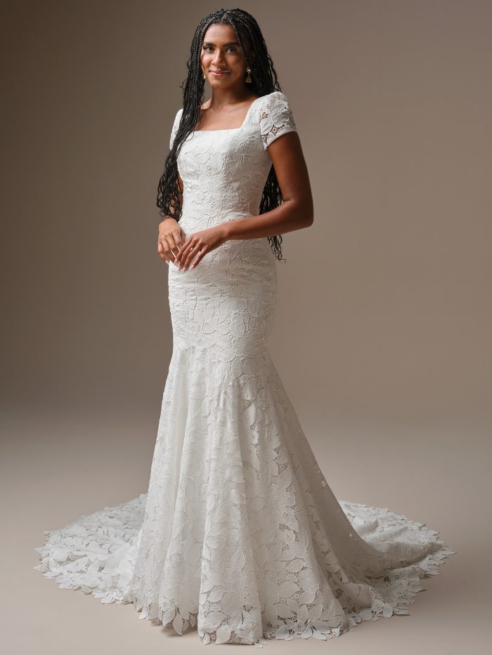 Dakota Leigh by Maggie Sottero, modest allover lace sheath wedding dress with square neckline, cap sleeves, covered buttons, and extended illusion lace train.