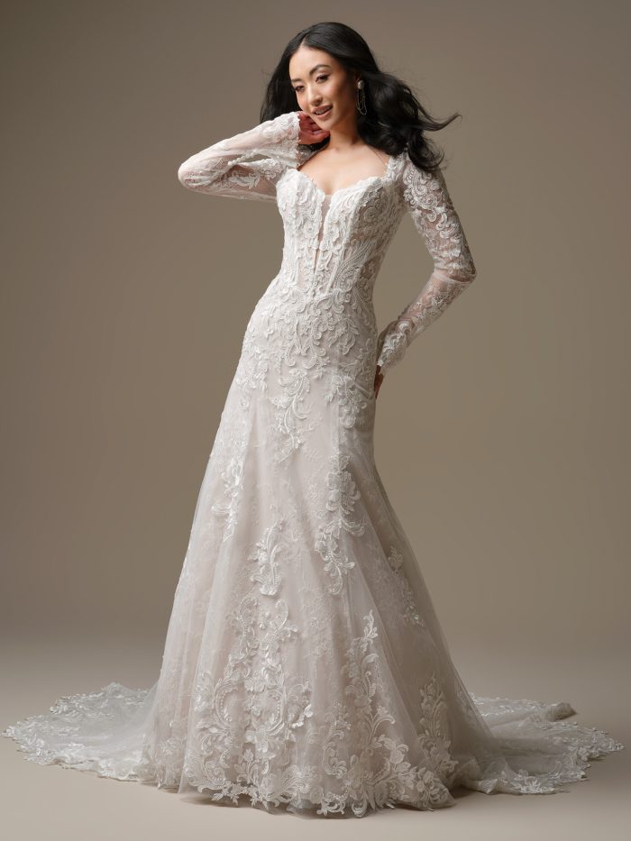 Farrah wedding dress featuring sequin lace motifs layered over Chantilly lace, an illusion lace bodice with exposed boning detail, a plunging illusion V neckline, a fitted silhouette that softly flares into an extended scalloped illusion lace train, and Chantilly lace covered buttons along the back. Model stands in a neutral studio setting.