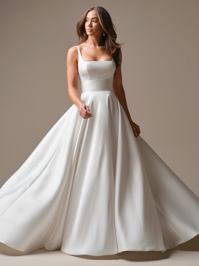 Galina by Maggie Sottero — a modern square-neck satin A-line wedding dress with wide straps and a flowing full skirt, styled with soft waves and minimal jewelry in a neutral studio setting.