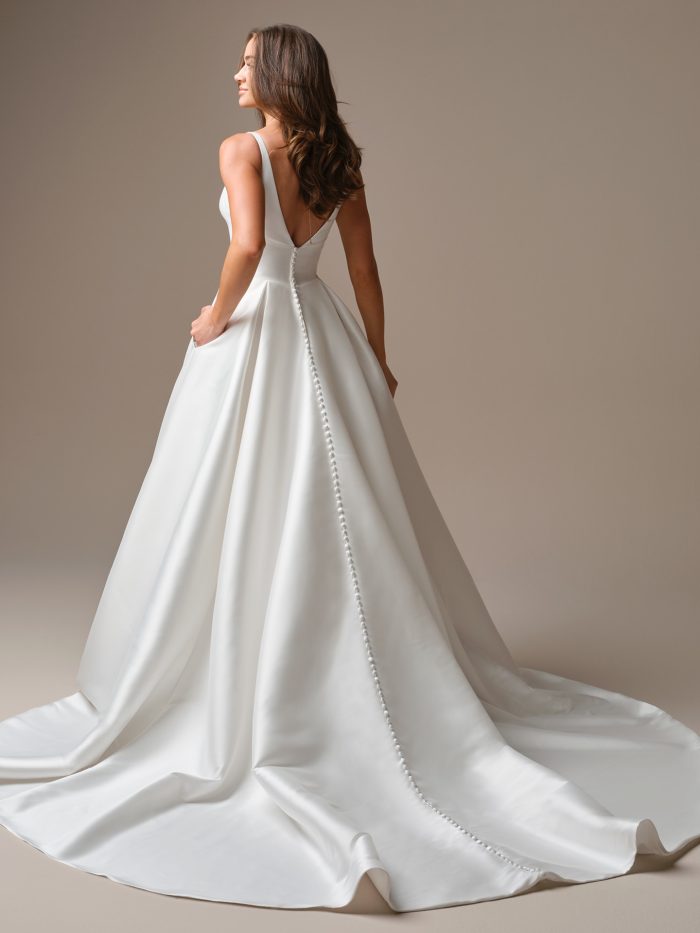 Galina by Maggie Sottero — back view of a square-neck satin A-line wedding dress featuring a deep V-back, full satin train, and buttons trailing down the skirt, styled with loose waves in a minimalist studio setting.