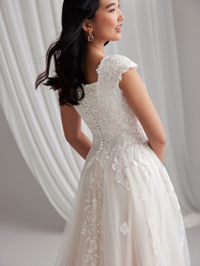 Back view of Maggie Sottero Harlem Leigh, showcasing the modest square back, cap sleeves, covered button-over-zipper closure, and lace motifs trailing onto the skirt and train.