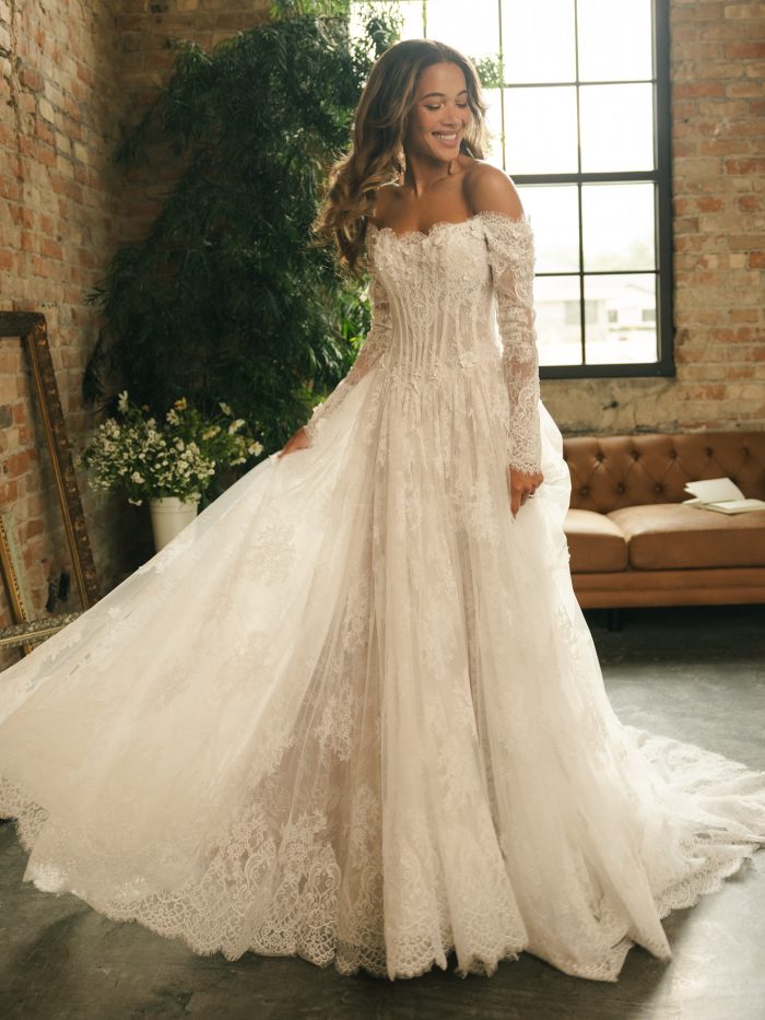 Model wearing the Kitaro ball gown wedding dress, featuring an off-the-shoulder lace bodice with floral appliqués, long illusion lace sleeves, and a full layered lace skirt for a romantic, vintage-inspired bridal look.
