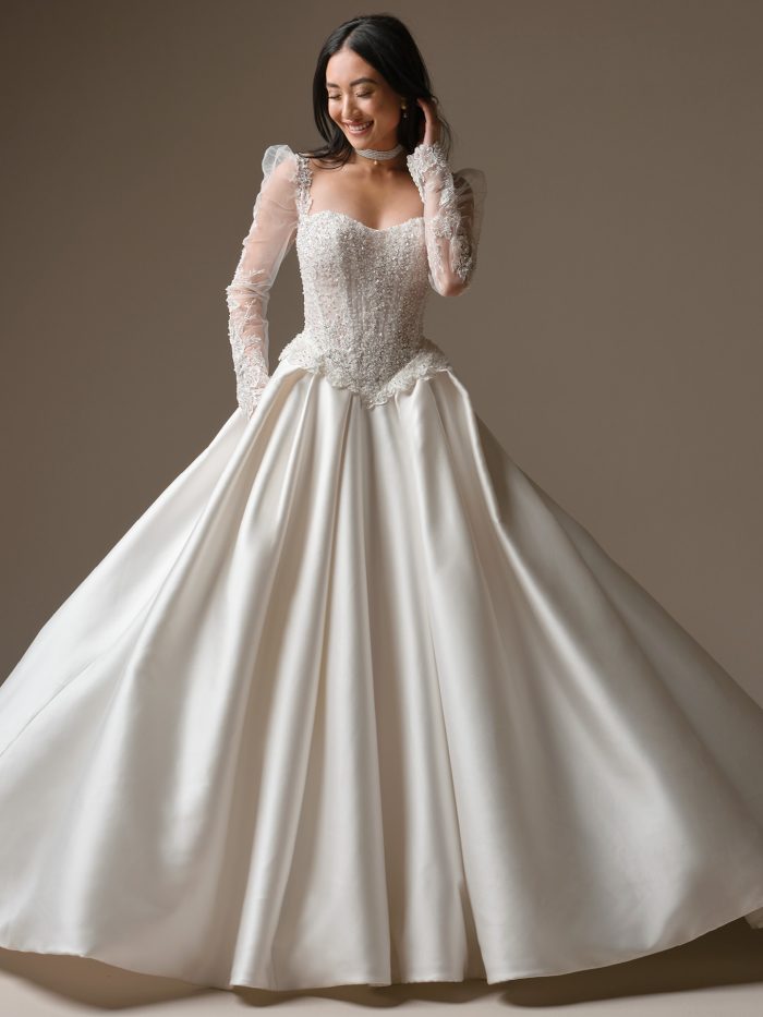 London wedding dress featuring a sheer organza-lined bodice with beaded lace motifs, long illusion sleeves, a soft sweetheart neckline, illusion lace tiered peplums at the hips, and a full Cartago satin ballgown skirt with pockets. Model stands in a studio setting while holding the skirt.