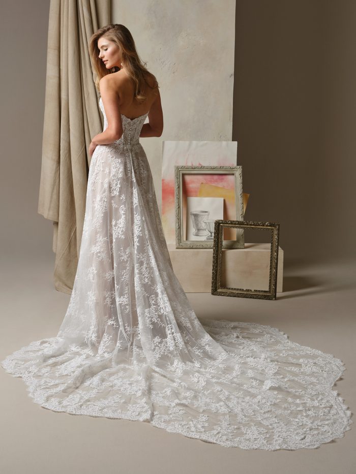 Back view of the Maggie Sottero Lucca A-line wedding dress showcasing a strapless lace bodice, flowing lace-overlay skirt, and extended scalloped lace train in a studio setting with framed artwork.
