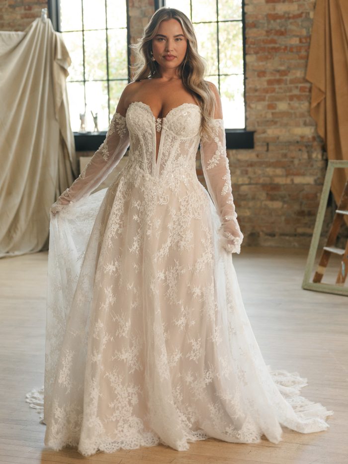 Lucca wedding dress featuring beaded and sequin lace motifs over Chantilly lace and glitter tulle, an illusion bodice with exposed boning detail, a deep illusion sweetheart neckline with lace medallion detail, and a soft A-line skirt with an extended lace train. Model stands in a softly lit loft-style studio.