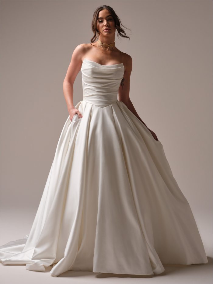Maureen by Maggie Sottero — timeless satin ballgown wedding dress featuring a softly draped cowl neckline, fitted bodice, and voluminous pleated skirt with pockets.