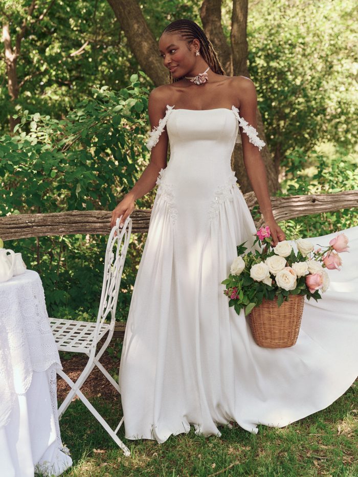 Bride wearing the Morwenna A-line wedding dress by Maggie Sottero, featuring a strapless satin bodice, soft off-the-shoulder floral straps, and a flowing skirt styled for an outdoor boho garden setting.