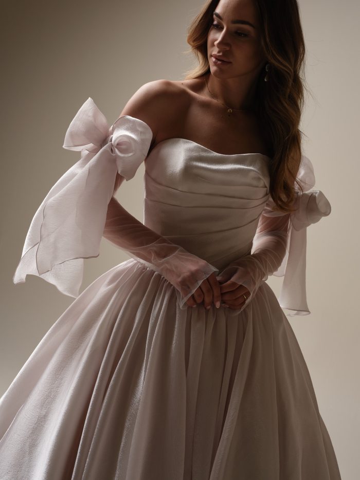 Paris by Maggie Sottero — close-up of a strapless ruched bodice with detachable sheer organza bow sleeves and pleated full skirt, styled with soft waves in a studio setting.