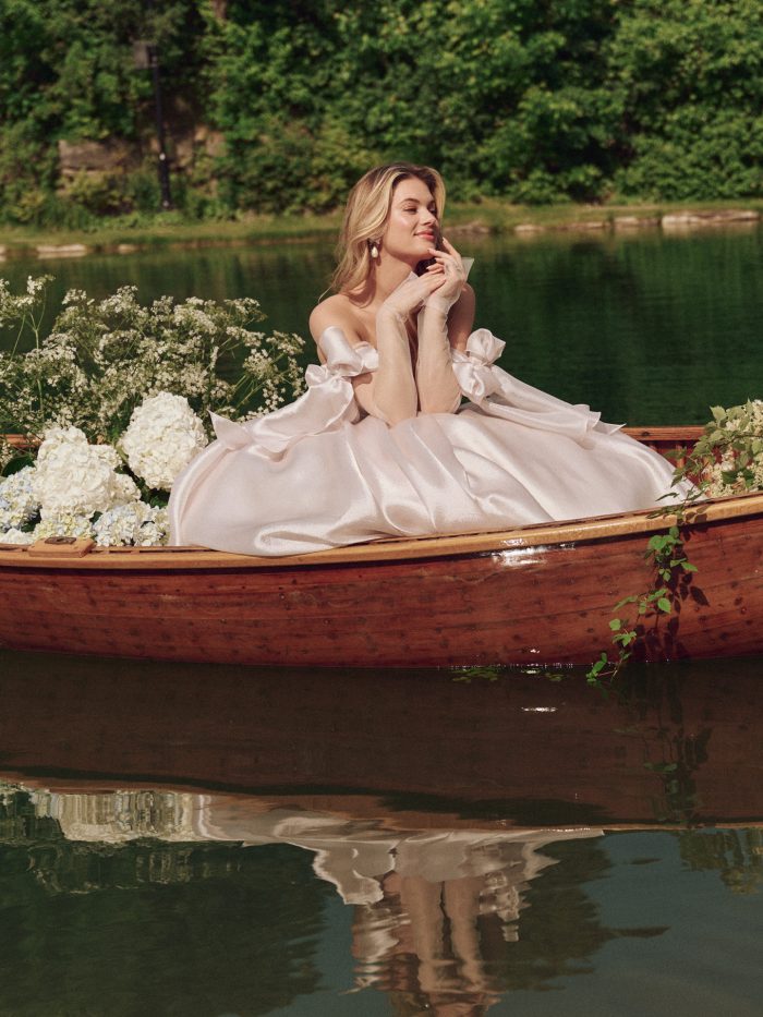 Bride wearing the Maggie Sottero Paris ballgown wedding dress in soft blush satin, seated in a wooden canoe on a lake, featuring an off-the-shoulder neckline with romantic bow details and voluminous skirt styled with sheer gloves and white florals.