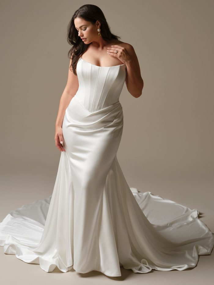 Model wearing Sinclaire by Maggie Sottero, a strapless satin fit-and-flare wedding dress with basque waistline, corset bodice, and smooth skirt.