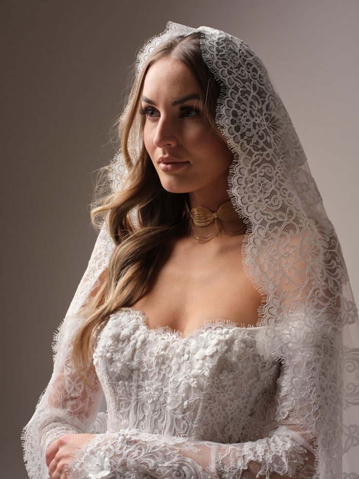 Bride wearing the Takera veil by Maggie Sottero, a royal-length mantilla veil with intricate Chantilly lace and scalloped edging framing her face and shoulders.