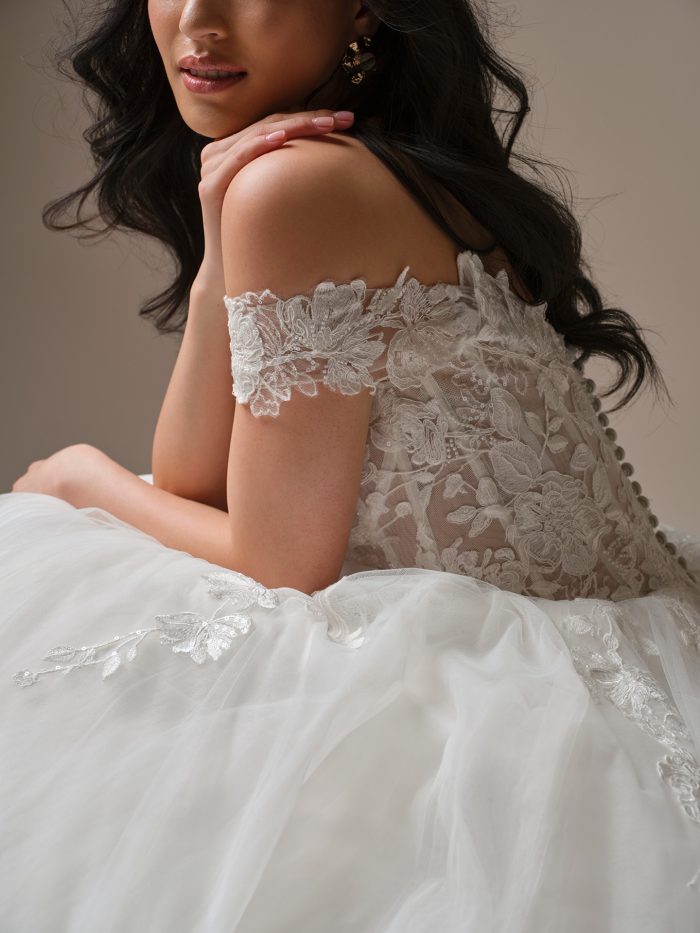 Close-up of the Verina wedding dress by Maggie Sottero highlighting the off-the-shoulder lace sleeves, illusion lace back with covered buttons, and delicate floral appliqué details.