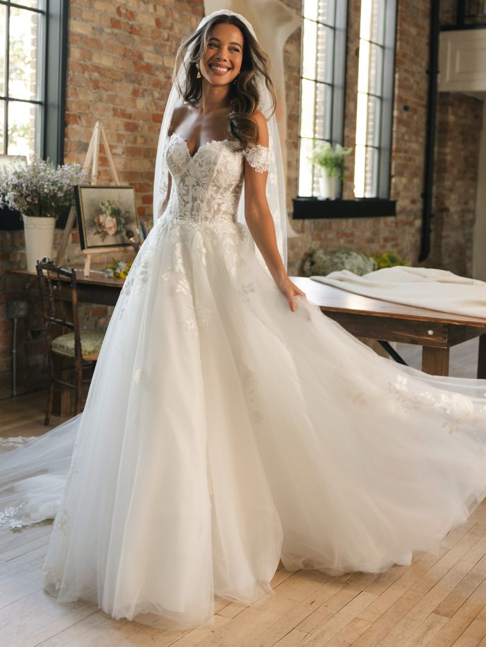 Verina by Maggie Sottero ball gown wedding dress featuring beaded and sequin corded lace over soft tulle with off-the-shoulder cap sleeves and a flowing skirt