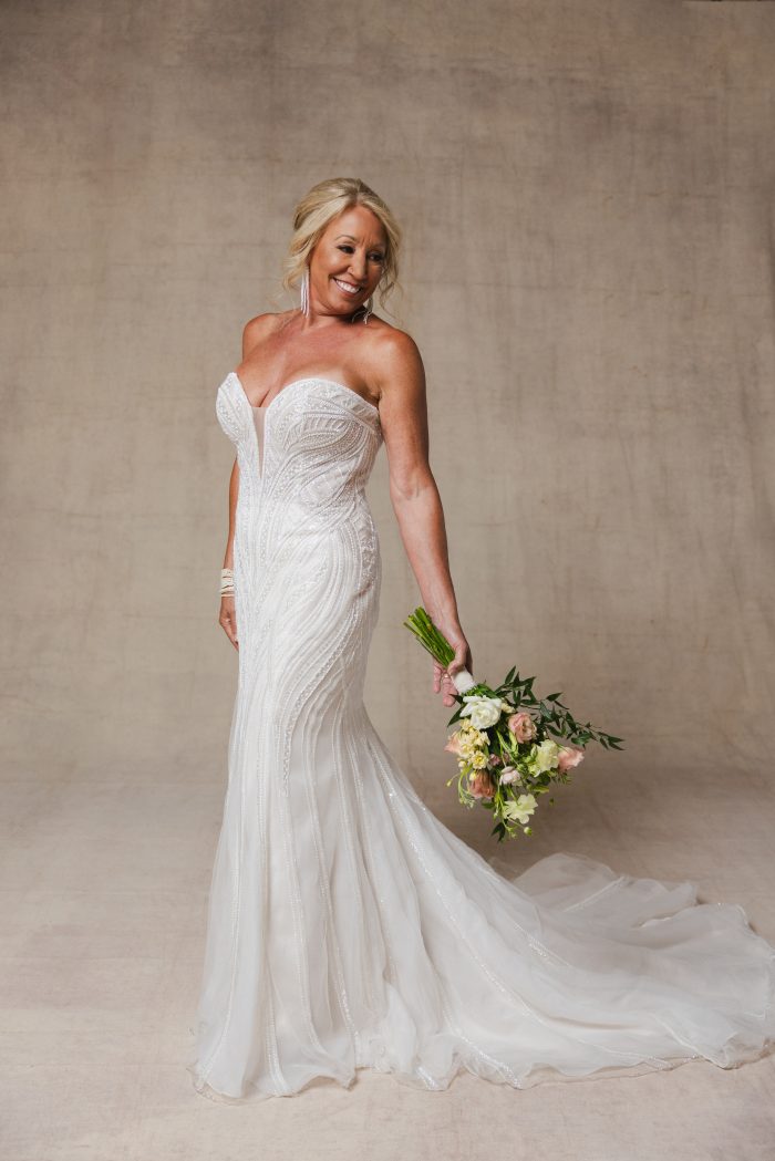 Older bride wearing a beaded fit and flare wedding dress holding a floral bouquet behind her