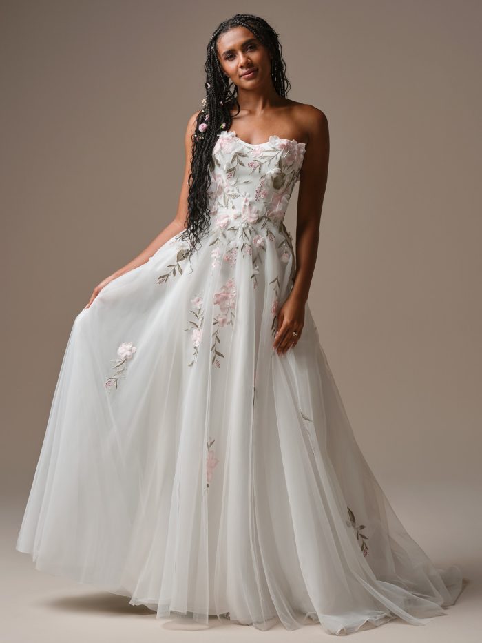 Front view of the Blossom wedding dress featuring a strapless bodice and airy A-line skirt with pink and white 3D floral appliqué and embroidered leaves.