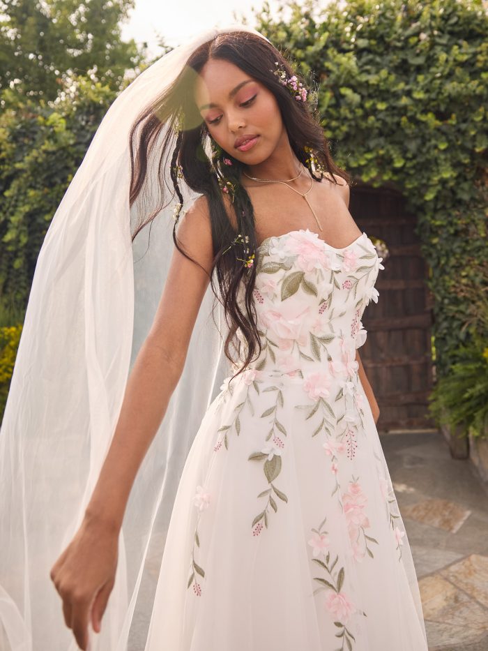 Close-up of the Blossom wedding gown by Rebecca Ingram, highlighting the strapless sweetheart neckline, pink floral appliqués, and leafy embroidered details, with a flowing veil and soft garden backdrop.