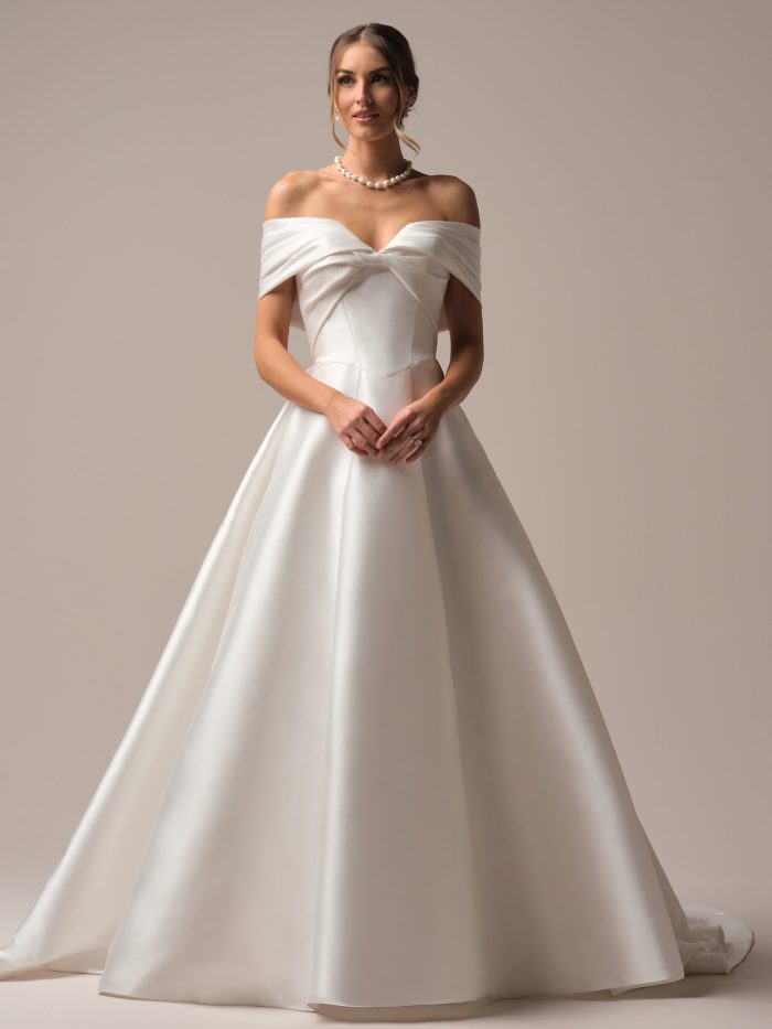 Model wearing the Rebecca Ingram Brooke A-line wedding dress with an off-the-shoulder Mikado bodice, strapless sweetheart neckline, and full skirt with pockets.