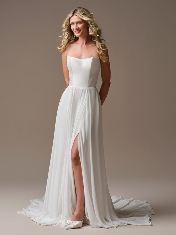 Model wearing the Rebecca Ingram Georgina strapless wedding dress with a cat-eye neckline, flowing chiffon skirt, and high side slit.