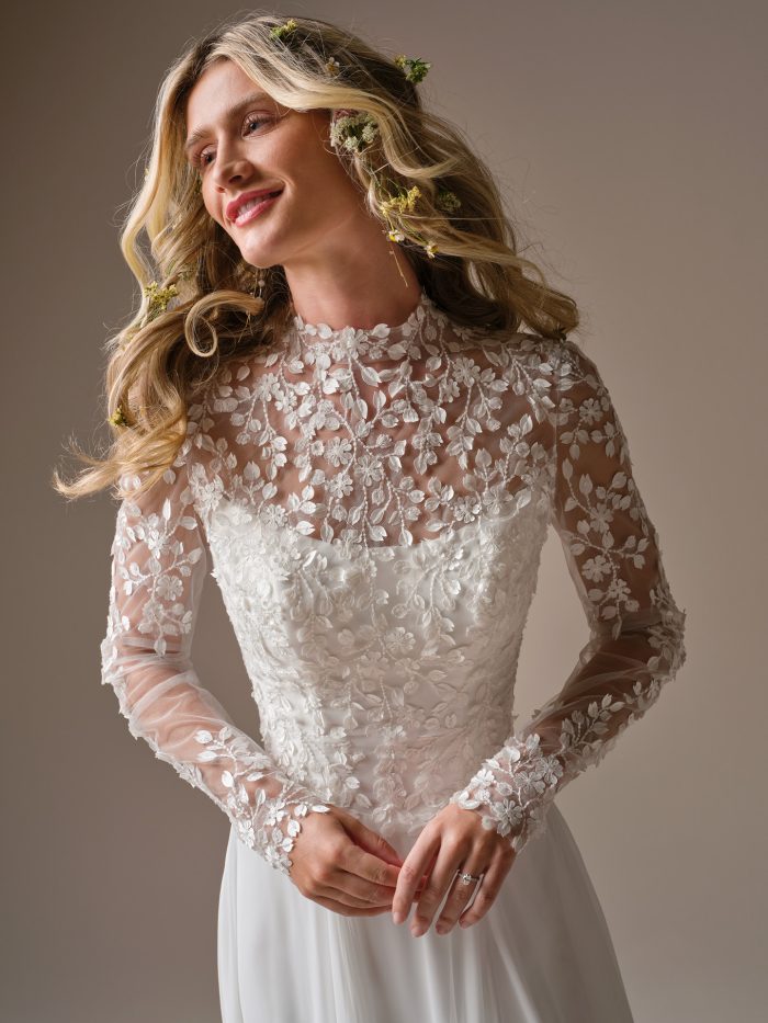 Model wearing the Rebecca Ingram Georgina wedding dress styled with a detachable illusion lace jacket featuring long sleeves and floral embroidery.