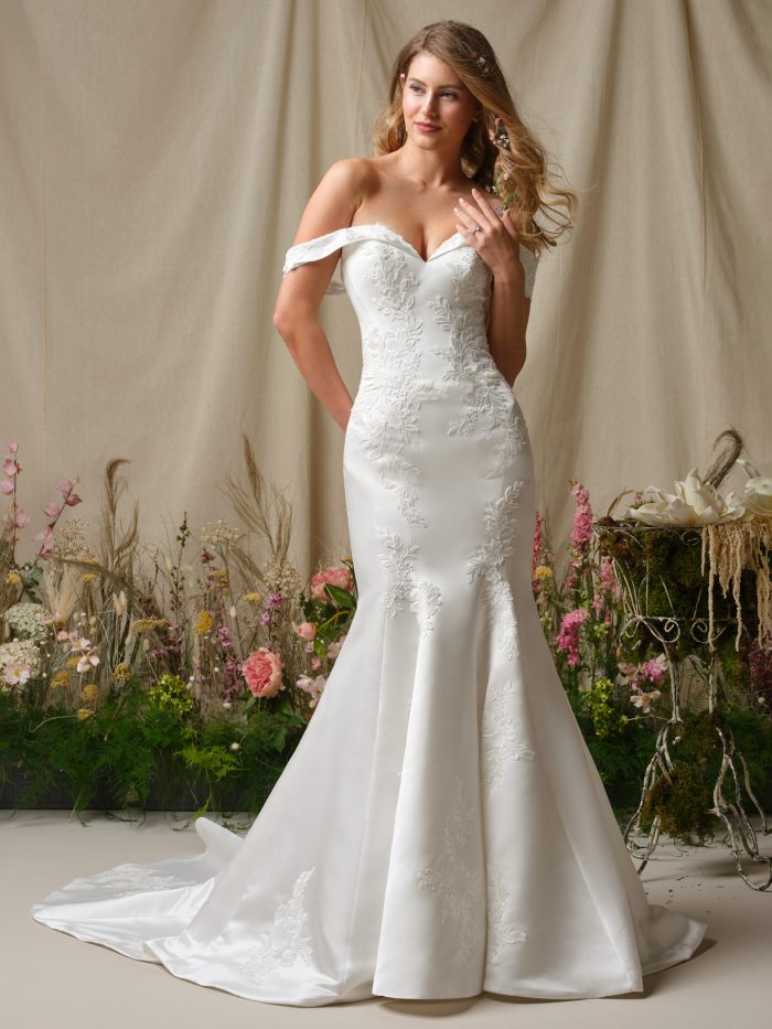Model wearing the Rebecca Ingram Miley fit-and-flare wedding dress with an off-the-shoulder portrait neckline, sequined lace accents, and a smooth satin skirt.