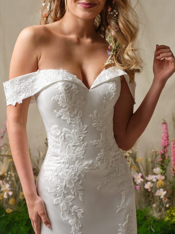 Close-up of the Rebecca Ingram Miley wedding dress highlighting the off-the-shoulder lace sleeves, portrait neckline, and sequined lace motifs on the bodice.