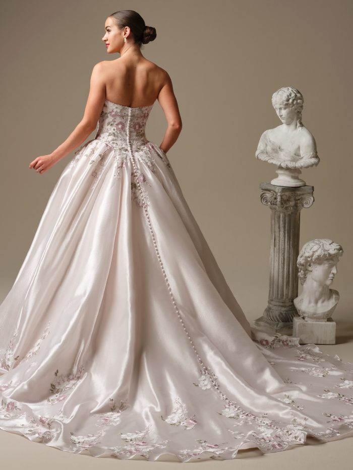 Back view of the Sottero and Midgley Alvarez ballgown wedding dress showcasing a strapless floral lace bodice with covered button closure, full satin skirt with scattered pink floral embroidery, and extended train styled beside classical marble busts.