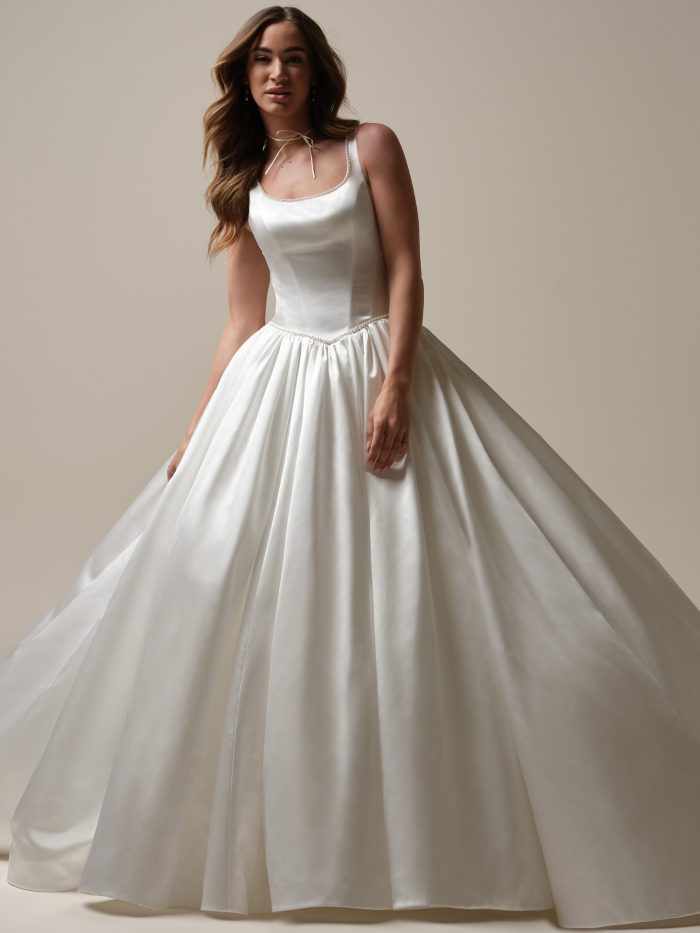 Bride wearing a satin ball gown wedding dress with a scoop neckline, pearl trim, and fitted bodice, posing gracefully in soft studio lighting.