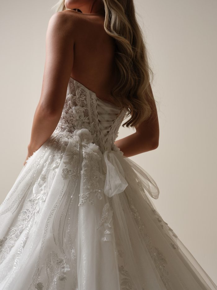 Back view of a strapless lace A-line wedding dress featuring a lace-up corset back, sheer bodice, 3D floral appliqués, and soft tulle bow detail at the waist.
