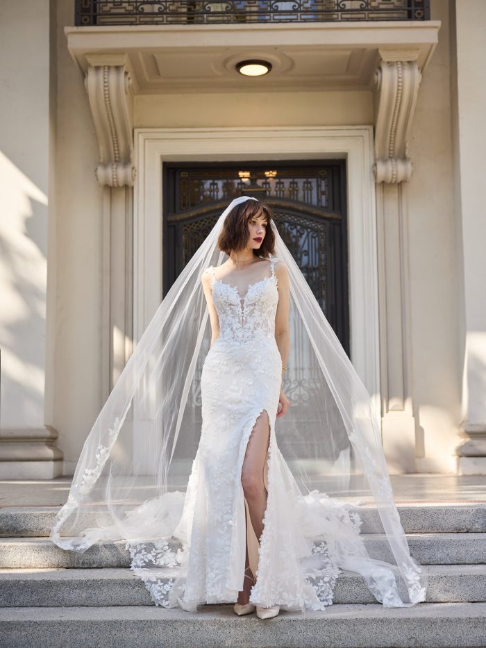 Bride wearing the West wedding dress, a fitted floral lace fit-and-flare gown with a thigh-high slit and deep illusion neckline, styled with a long cathedral veil while standing on marble steps outside an ornate building.