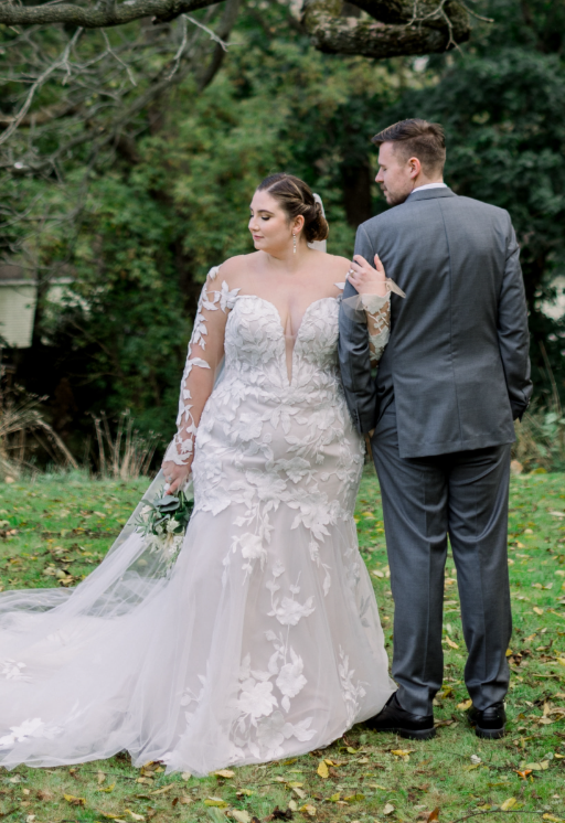 Bride in a long-sleeve lace wedding dress with plunging neckline, 3D floral appliqués, and chapel train, holding hands with groom in a gray suit during outdoor portraits.
