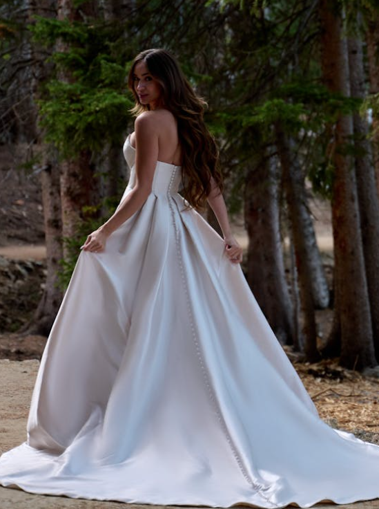 Back view of strapless satin ball gown wedding dress featuring covered buttons down the train, styled in a forest setting for a romantic bridal look.